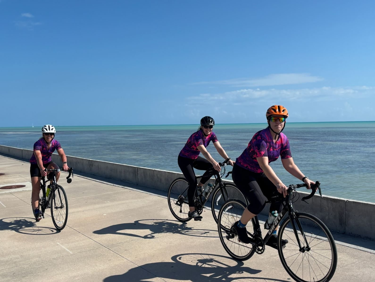 Three riders in matching jerseys cycling a seawall path beside turquoise water in the Florida Keys