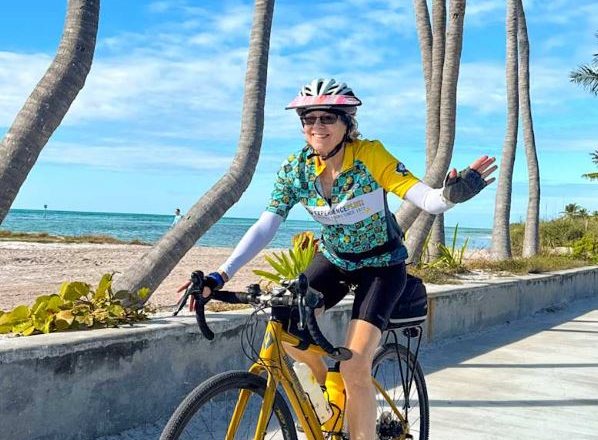 A rider waving from her bike on a palm-lined waterfront path