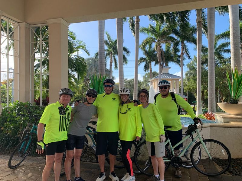 Six riders in bright yellow shirts with their bikes under palm trees
